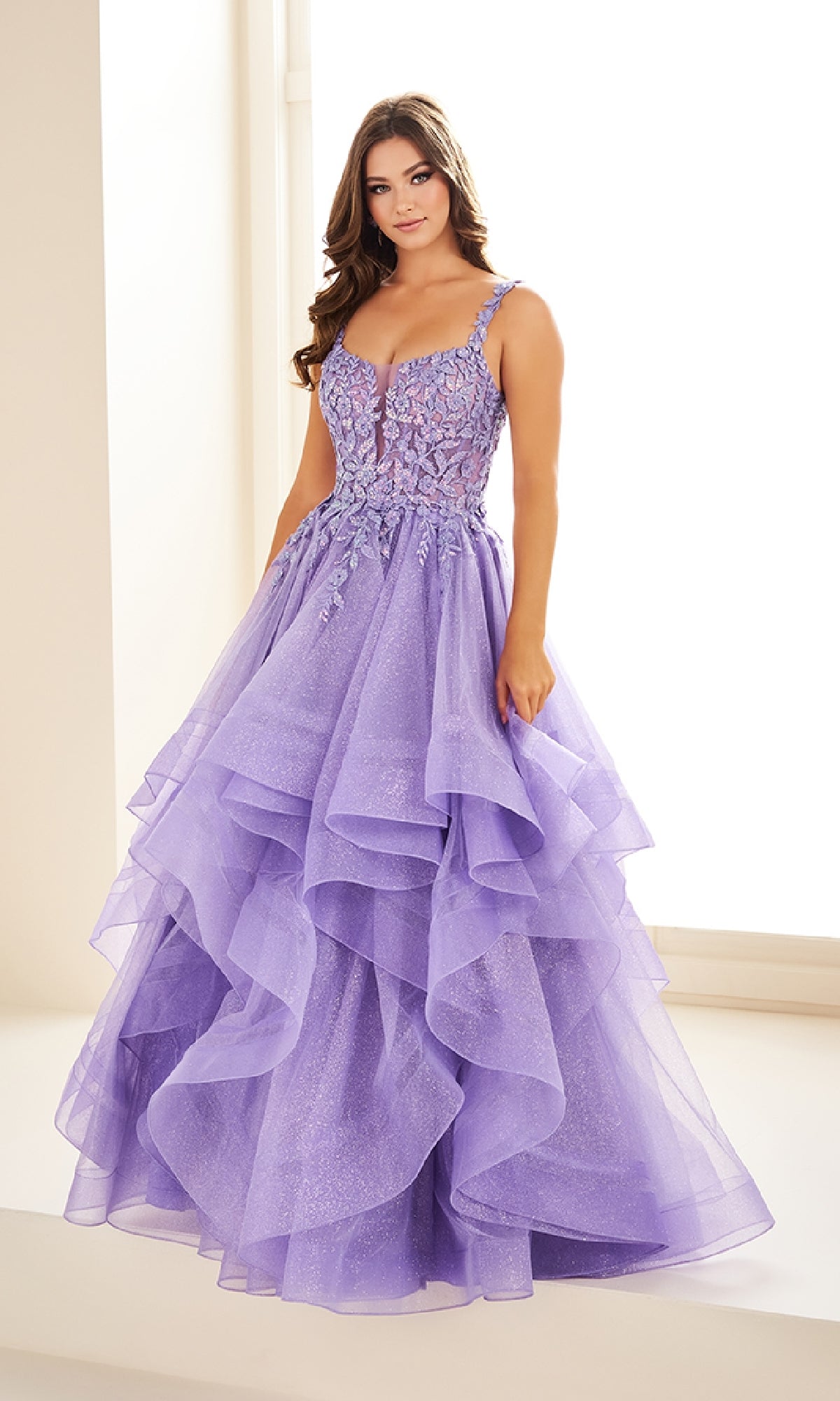 A woman stands indoors near a bright window wearing the Ellie Wilde EW36035, a long glitter prom ball gown.